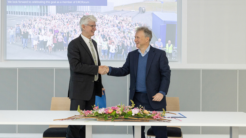 Photograph of two men shaking hands in front of a screen. A table in front of them holds flowers and an open folder.